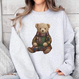 May include: A light gray sweatshirt featuring a seated teddy bear illustration. The bear wears a plaid scarf, a green sweater, and plaid pants. The bear's fur is light brown, and the overall design has a cozy, autumnal feel.