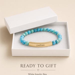 May include: A stretch bracelet featuring round turquoise-colored beads with natural veining and a central curved gold-tone metal bar engraved with the word custom. The bracelet is presented inside an open white rectangular jewelry box.