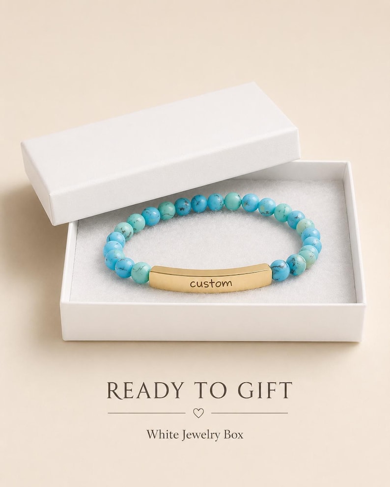 May include: A stretch bracelet featuring round turquoise-colored beads with natural veining and a central curved gold-tone metal bar engraved with the word custom. The bracelet is presented inside an open white rectangular jewelry box.