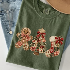 May include: An olive green t-shirt featuring a graphic print of a gingerbread person, a Christmas tree, and a stocking. Each figure is decorated with red and white plaid patterns, green holly leaves, and red bows. The shirt has a Comfort Colors label.