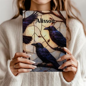 May include: A notebook with a stained-glass design featuring three black ravens perched on branches. The notebook cover has the name "Alisson" in elegant script. The background has a soft, yellow glow.