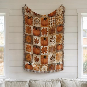 May include: A woven throw blanket featuring a grid pattern of orange pumpkins and various autumn leaves in shades of brown, tan, and cream. The rectangular textile has a fringed edge along the bottom and is displayed hanging vertically.