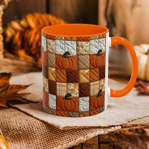 May include: A ceramic mug featuring a patchwork quilt pattern with alternating squares of orange pumpkins, brown plaid, and cream-colored leaves. The mug has a bright orange interior and a matching orange handle, with a quilted texture design.