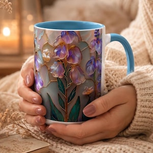 May include: A ceramic mug featuring a stained-glass style illustration of purple and white flowers with green leaves. The mug has a light blue interior and a matching light blue handle, with a white exterior base and gold-toned outlines.