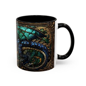 May include: A ceramic mug with a black handle and interior, featuring a wrap-around design of a blue and teal dragon with intricate scales. The dragon is set against a dark, ornate gold-toned frame that resembles stained glass artwork.