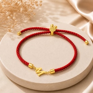 May include: A red braided bracelet with gold-coloured accents and an initial charm. The bracelet has a sliding knot closure for adjustable sizing. The initial charm is a stylized letter "M".