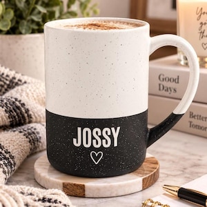 May include: A two-toned ceramic mug with a speckled finish. The top half is white and the bottom half is black. The word JOSSY is printed in white capital letters on the black section, followed by a small white heart outline below the text.