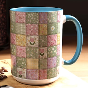 May include: A white ceramic mug with a patchwork design featuring floral patterns in pink, green, and yellow squares. The mug has a light blue interior and handle, perfect for enjoying coffee or tea.