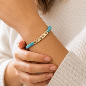 May include: A beaded bracelet featuring round, bright blue turquoise-colored beads and a central rectangular gold-toned metal bar. The bar is engraved with the name Charlie and a small paw print icon on each side of the text.