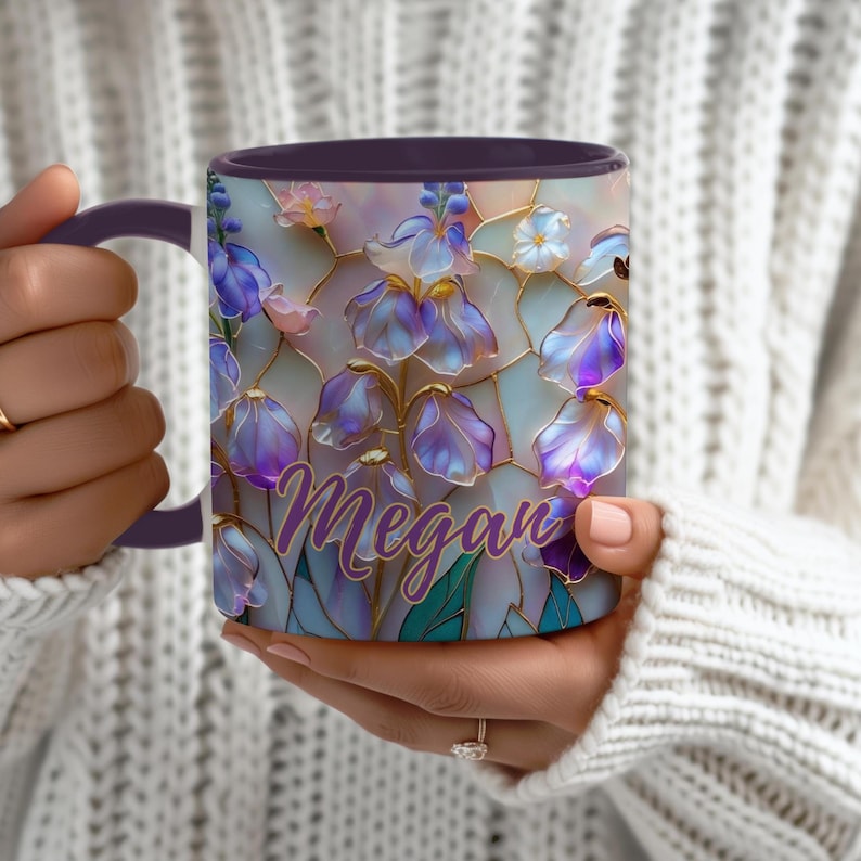 May include: A ceramic mug with a dark purple handle and rim, featuring a stained-glass style floral pattern in shades of blue, purple, and white. The name Megan is printed in a cursive purple font across the center of the floral design.