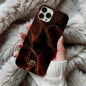 May include: A brown and black phone case with a swirling design and gold initials "JR". The case has a glossy finish and is held in a person's hand. The phone case has a camera bump with three lenses.