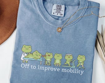 May include: A blue jean colored t-shirt featuring a graphic of six green frogs engaged in physical therapy activities. Below the frogs, the white text reads, "Off to improve mobility." The shirt has a crew neckline and a Comfort Colors brand label.