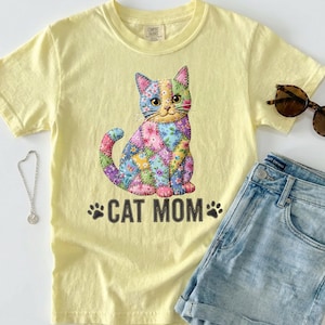May include: A light yellow t-shirt featuring a colorful patchwork cat design with the text "CAT MOM" in black. The cat design is made up of various floral and patterned fabric pieces. The shirt is paired with denim shorts and sunglasses.