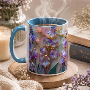 May include: A ceramic mug with a blue handle and rim, featuring a stained-glass style floral pattern of purple and blue flowers with gold outlines. Steam rises from the top of the mug, which is filled with a hot beverage, showcasing a vibrant design.