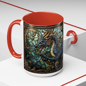 May include: A ceramic mug with a bright red handle and interior. The exterior features a detailed graphic of a blue dragon set against a background of teal and green stained glass patterns with ornate gold filigree borders and accents.