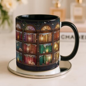 Peut inclure: Un mug en c&eacute;ramique avec une anse et un int&eacute;rieur noirs, orn&eacute; d'une impression panoramique d'une biblioth&egrave;que en bois &agrave; plusieurs niveaux remplie de livres color&eacute;s, de bougies allum&eacute;es, de petites fleurs blanches et de papillons.