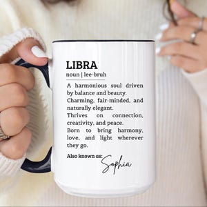 May include: A white ceramic mug with a dark blue handle and interior rim. The front features black printed text defining the word LIBRA as a noun, followed by a descriptive paragraph about the zodiac sign and the name Sophia in script font.