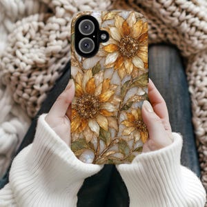 以下が含まれることがあります： 金色の花びら、暗い質感のある中心部、緑の葉が描かれた詳細なヒマワリ模様のスマートフォンケース。白い背景に光沢のあるステンドグラスのようなデザインで、黒いカメラレンズ用の切り抜きがあります。