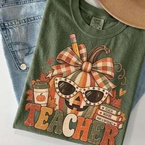May include: An olive green t-shirt featuring a graphic of a pumpkin wearing polka-dot sunglasses, topped with a plaid bow and a pencil. The design includes a coffee cup, autumn leaves, and a stack of books labeled TEACH, INSPIRE, MAKE A DIFFERENCE.
