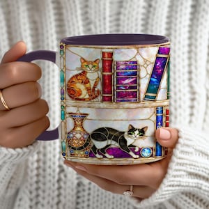 May include: A ceramic mug with a dark purple interior and handle, featuring a stained-glass style design of two cats on shelves with books and a vase. The top shelf shows an orange tabby cat, while the bottom shelf displays a black and white cat.