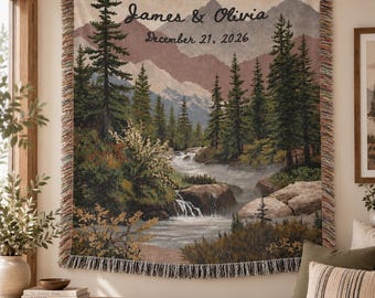 May include: A woven tapestry throw blanket featuring a landscape of mountains, pine trees, and a flowing stream. The text "James & Olivia" and "December 21, 2026" is printed at the top. The blanket has a fringed edge with multicolored threads.
