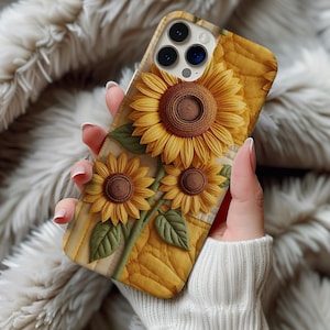 May include: A yellow and brown sunflower phone case featuring three sunflowers with green leaves. The case has a textured, quilted background. The phone case is held in a person's hand.