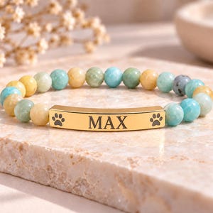 May include: A beaded bracelet featuring alternating light blue, yellow, and green polished stones. A central gold-toned metal bar is engraved with the name MAX, flanked by two small black paw print icons on either side of the text.