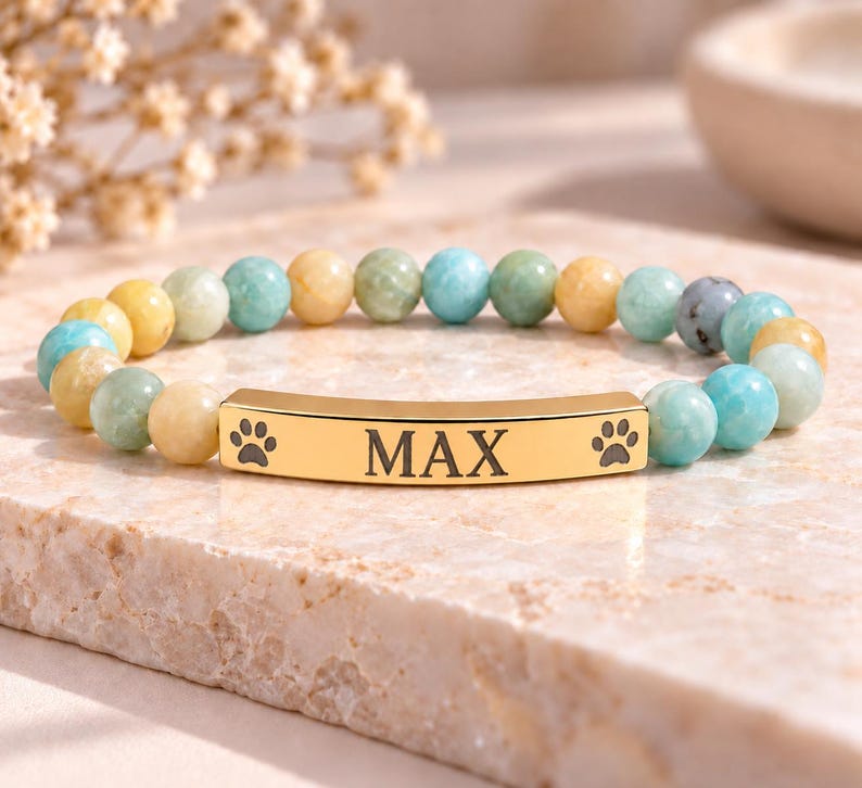 May include: A beaded bracelet featuring alternating light blue, yellow, and green polished stones. A central gold-toned metal bar is engraved with the name MAX, flanked by two small black paw print icons on either side of the text.