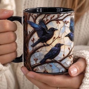 May include: A ceramic mug with a black handle and interior, featuring a stained-glass style design of three black crows perched on brown tree branches against a background of iridescent, light-colored, segmented glass panels.