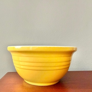 May include: A bright yellow ceramic mixing bowl with horizontal ridged detailing. The bowl has a wide rim and a rounded shape, perfect for kitchen use. The bowl is sitting on a wooden surface.