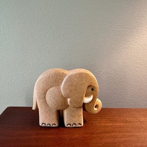 May include: A tan elephant figurine with a rounded body, large ears, and a curled trunk. The elephant has small black eyes and white tusks. The figurine is on a wooden surface.