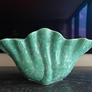 May include: A seafoam green ceramic seashell-shaped bowl with white speckled accents. The bowl has a scalloped edge and a square base. The bowl is likely used for decorative purposes or as a serving dish.