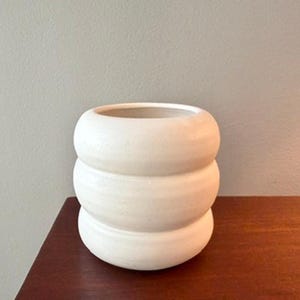 May include: A cream-coloured ceramic vase with a stacked, rounded design. The vase has a wide opening and sits on a dark wooden surface. The neutral colour and unique shape make it a decorative accent piece.