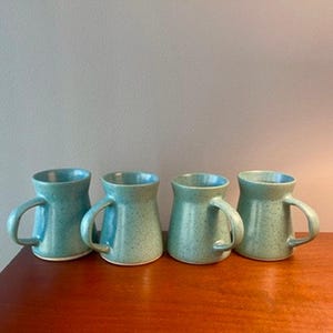 Może przedstawiać: Cztery turkusowe ceramiczne kubki z nakrapianymi detalami. Każdy kubek ma zaokrąglony korpus, zakrzywiony uchwyt i jasne wnętrze. Kubki są ułożone na drewnianej powierzchni.