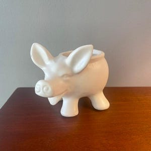 Peut inclure: Une figurine de cochon en céramique crème avec un dos creux. Le cochon a de grandes oreilles, un groin avec des narines et quatre pattes courtes. La figurine est posée sur une surface en bois brun foncé.