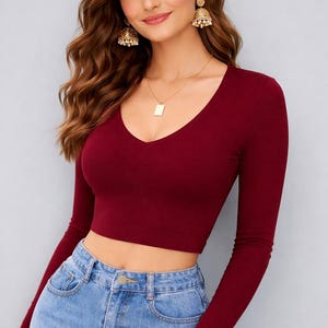 Op de afbeelding: Een bordeauxrode crop top met lange mouwen en een diepe V-hals wordt gecombineerd met lichtblauwe jeans. De top is nauwsluitend. Het model draagt gouden oorbellen en een ketting.