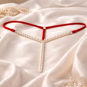 Könnte beinhalten: Ein roter String-Tanga mit Perlenbesatz. Das Kleidungsstück hat ein rotes Band und eine vertikale Perlenkette. Zusätzliche Perlen sind im Hintergrund auf weißem Satin-Stoff verstreut.