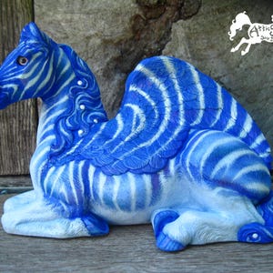 May include: A ceramic figurine of a winged creature with the head of a horse and the body of a camel, painted with blue and white zebra-like stripes. The figure features a flowing mane, feathered wings, and the text Attic Studio WL on the top right.