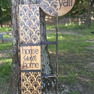 May include: A wooden porch sign set featuring a tall rectangular board with a black fan-like pattern and the text home sweet home, paired with a round sign displaying the text hey yall and a black bow with green foliage accents.