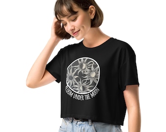 Puede incluir: Top corto negro con un diseño circular de flores grises estilizadas. El texto "BLOOM UNDER THE MOON" está impreso en blanco alrededor del círculo. El top se combina con unos vaqueros azules. Diseño sencillo y elegante.
