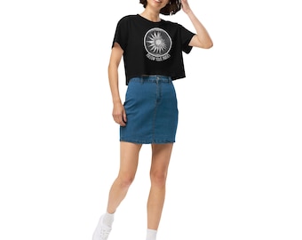 Puede incluir: Un top corto negro con un gráfico de sol blanco y el texto "FOLLOW YOUR PATHS". Se combina con una falda vaquera azul. La modelo lleva zapatillas y calcetines blancos.