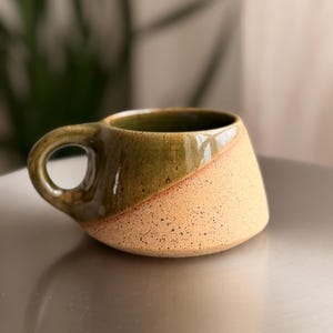 May include: A ceramic mug with a wide, tapered base and a circular handle. The upper portion features a glossy, dark olive green glaze, while the lower section is composed of a textured, speckled beige clay with a matte, natural finish.