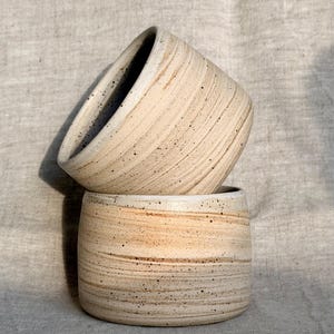 May include: Two handmade ceramic cups with a beige and brown striped design. The cups are stacked, with one slightly tilted, revealing a dark interior. The speckled texture and organic shape suggest a rustic, artisanal aesthetic.