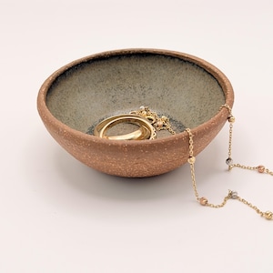 Può includere: Una piccola ciotola in ceramica fatta a mano con interno grigio maculato ed esterno marrone rossiccio. La ciotola contiene gioielli in oro, tra cui un anello e una collana di perline. Il portagioie è appoggiato su una superficie bianca.
