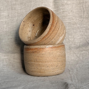 May include: Two handmade ceramic cups stacked on top of each other. The cups are a speckled beige color with a reddish-brown band around the rim. The cups have a rustic, earthy aesthetic.