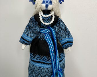 May include: A handmade doll with a white face and a blue and white cross design. The doll is wearing a black and blue dress with a blue sash and a white beaded necklace. The doll has long white hair and a wreath of blue and white flowers on her head.
