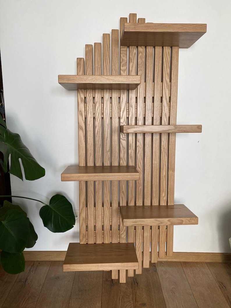 May include: A wooden wall-mounted shelf unit with multiple shelves and vertical slats. The shelves are rectangular and vary in size, offering a display space. The unit is made of light-colored wood, showcasing the natural grain. The design is modern and functional.