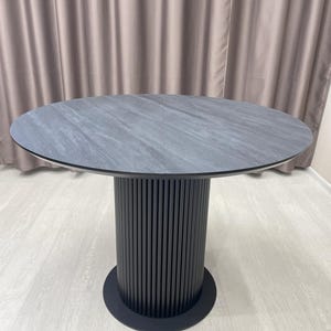 May include: A round dining table with a gray stone-like top and a black, vertically-slatted pedestal base. The table is approximately 48 inches in diameter and stands on a black circular base.
