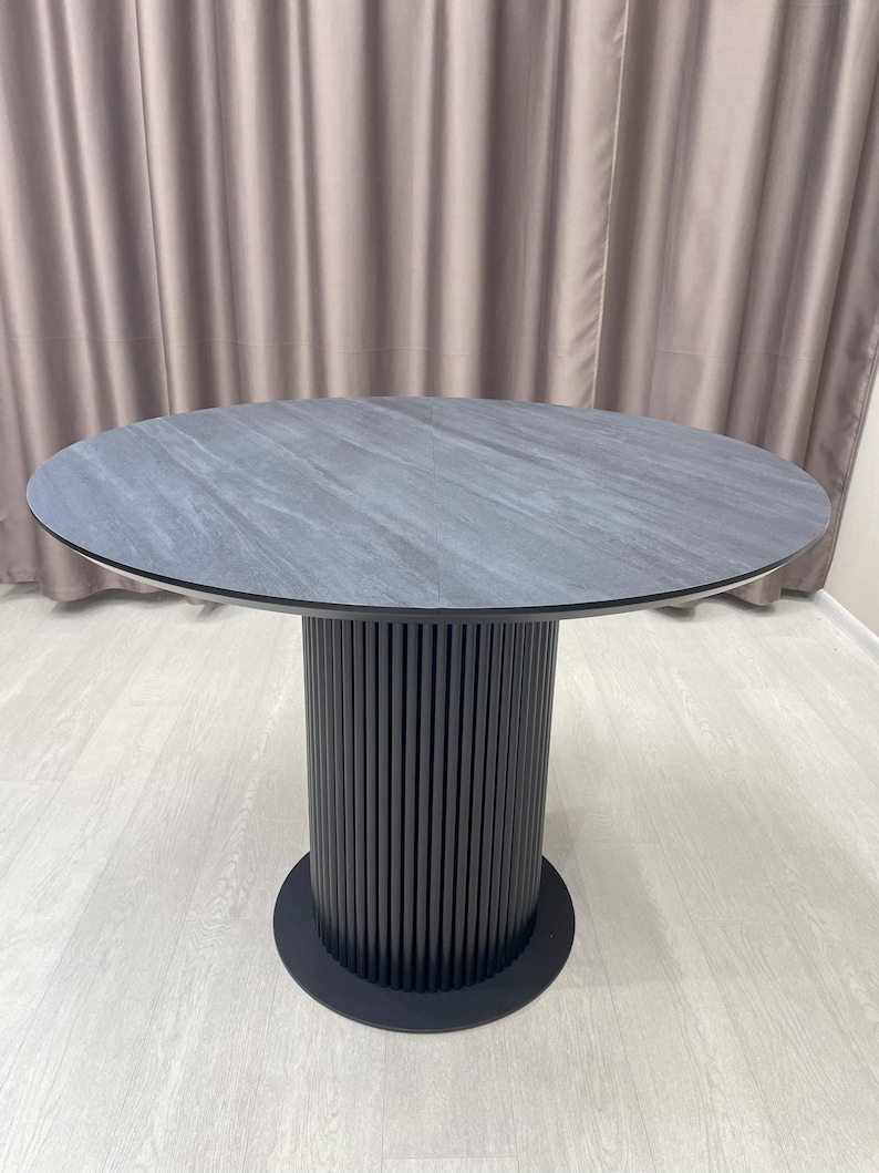 May include: A round dining table with a gray stone-like top and a black, vertically-slatted pedestal base. The table is approximately 48 inches in diameter and stands on a black circular base.