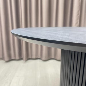 May include: A modern, round table with a gray, textured tabletop and a silver-colored edge. The table has a cylindrical base with vertical, gray, ridged detailing. The background features a neutral-colored curtain.
